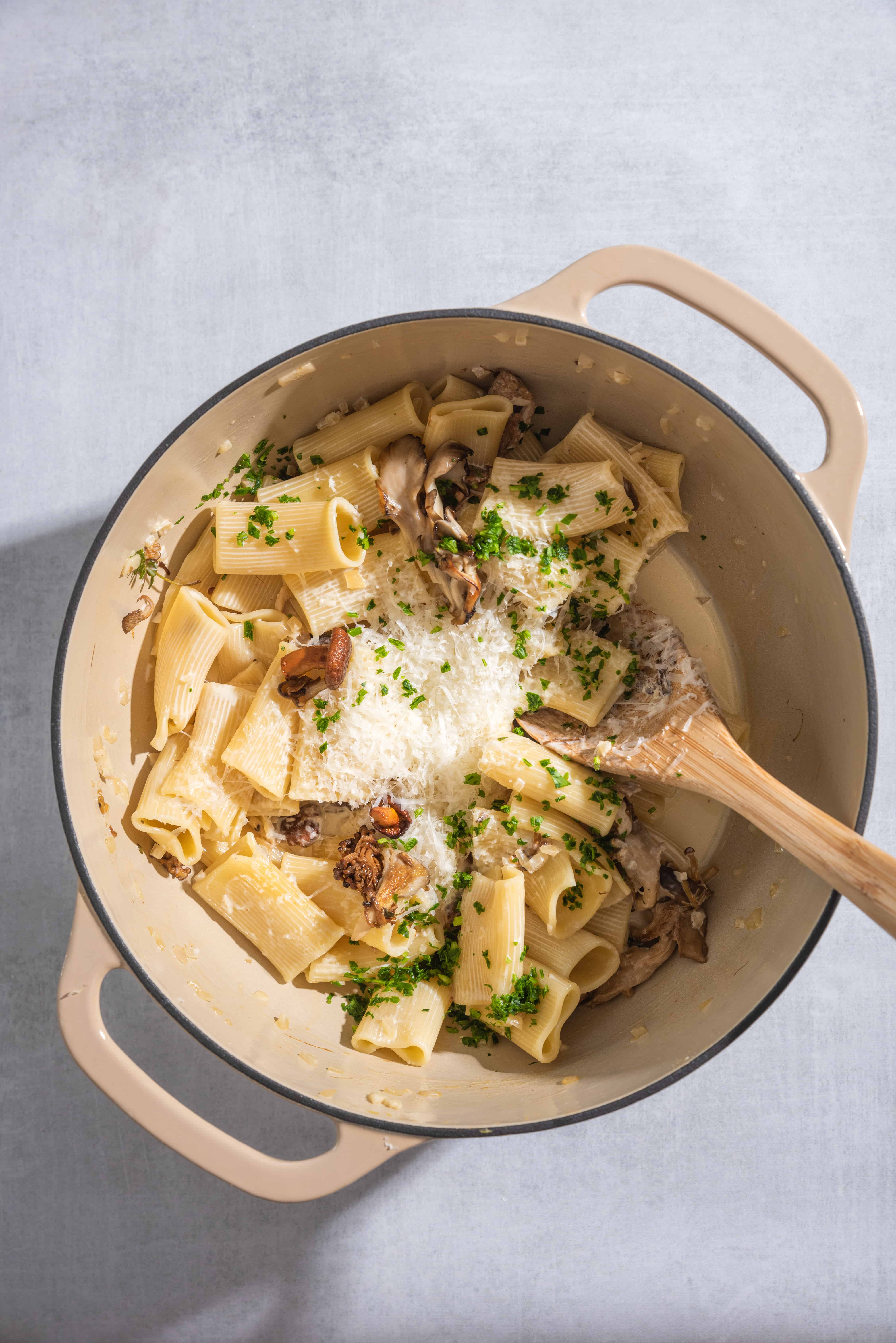 a beige dutch oven filled with rigatoni pasta, mushrooms, parmesan cheese and a wooden spoon