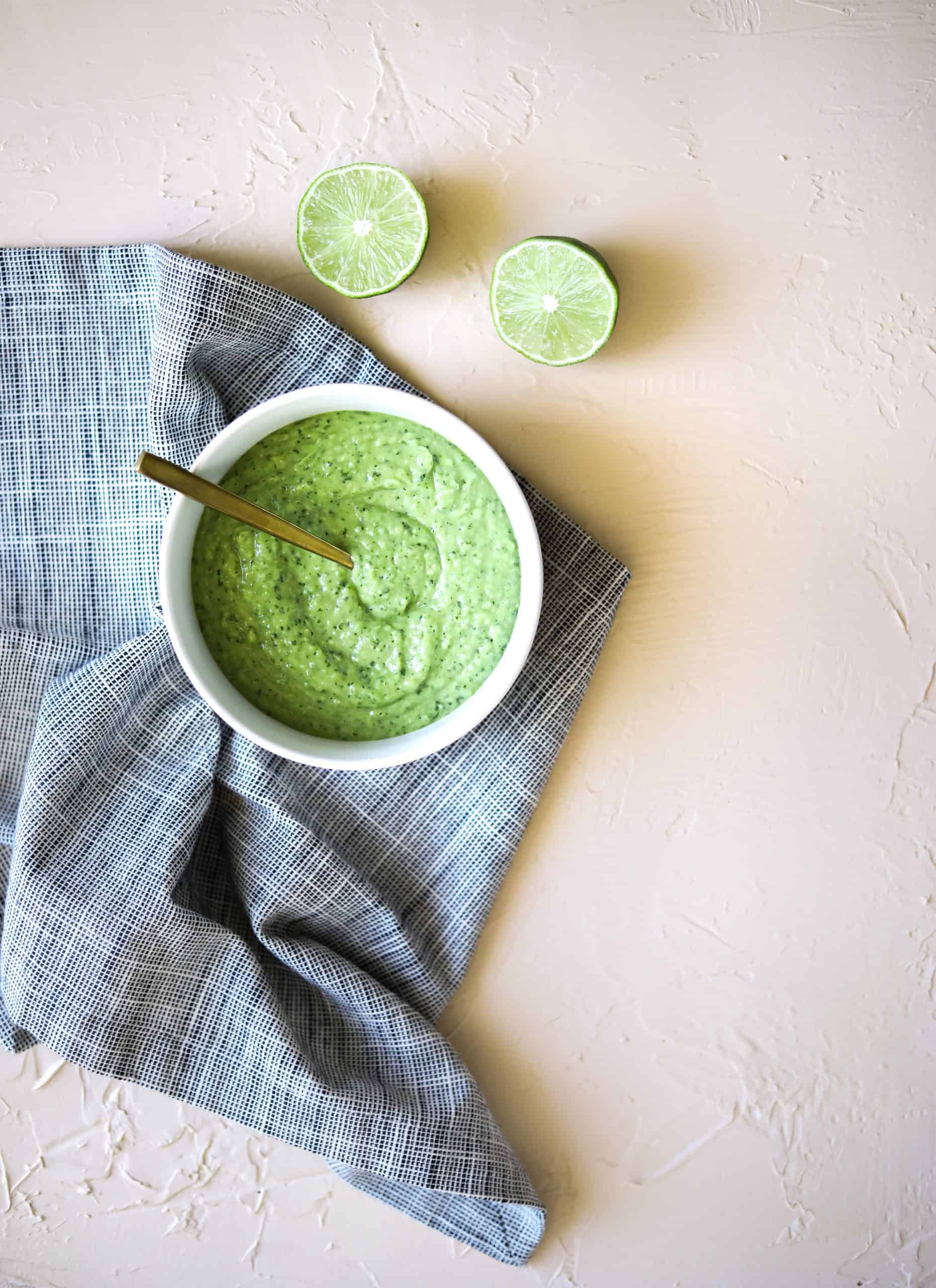 Creamy avocado salsa in a white bowl with a blue cloth and two limes on the side.