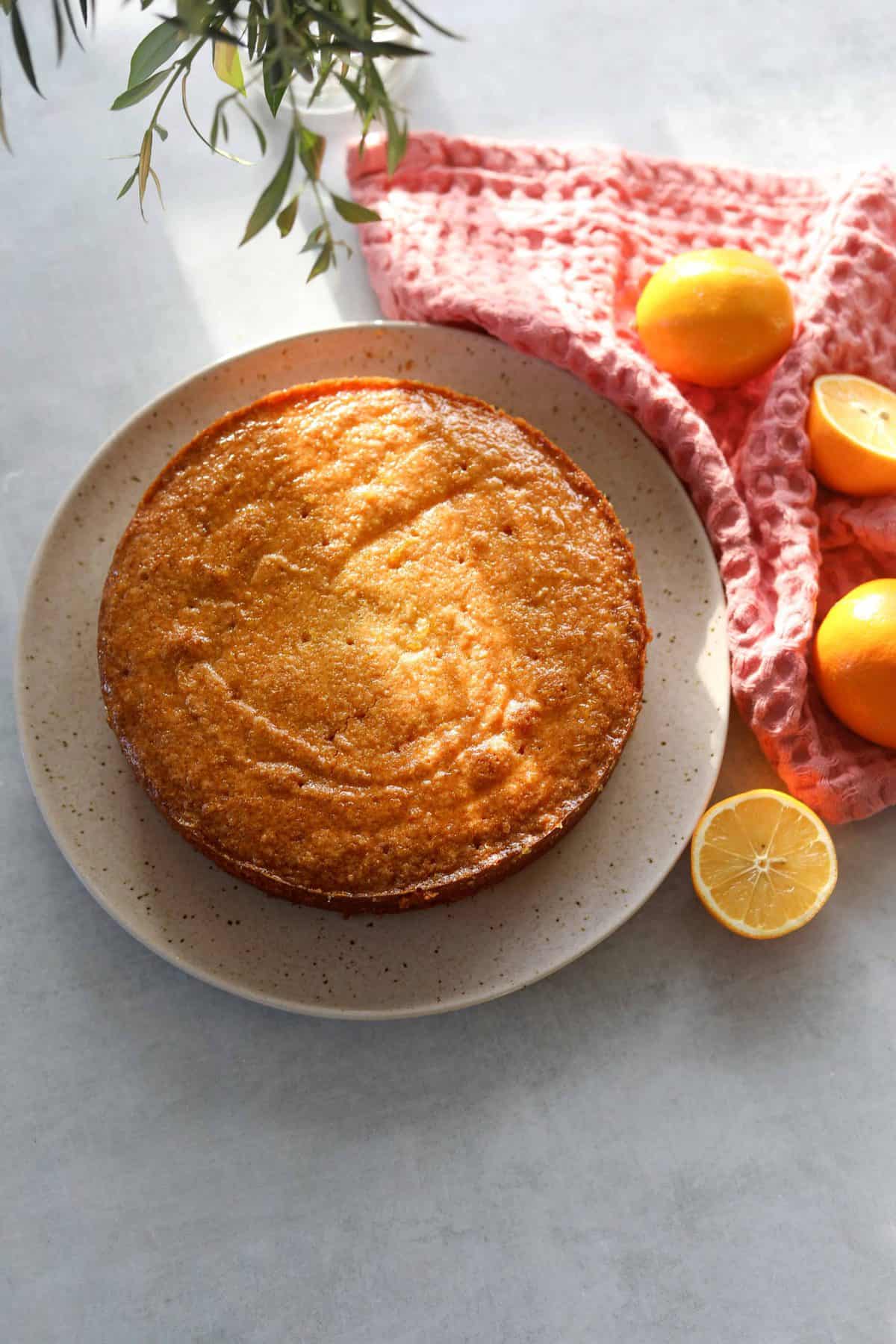 Meyer lemon polenta cake sitting on a cream plate surrounded by lemons and a pink tea towel