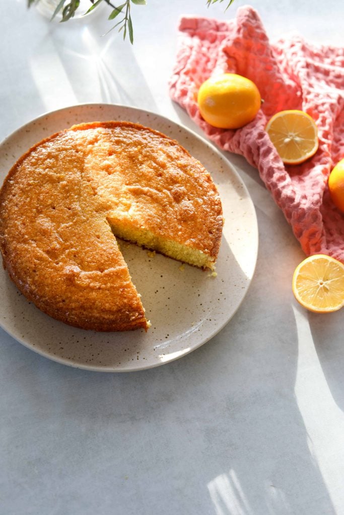 Meyer lemon polenta cake with a slice missing sitting on a cream plate surrounded by lemons and a pink tea towel