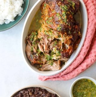 tender roasted pork in an oval baking dish with side dishes of rice, beans and mojo sauce