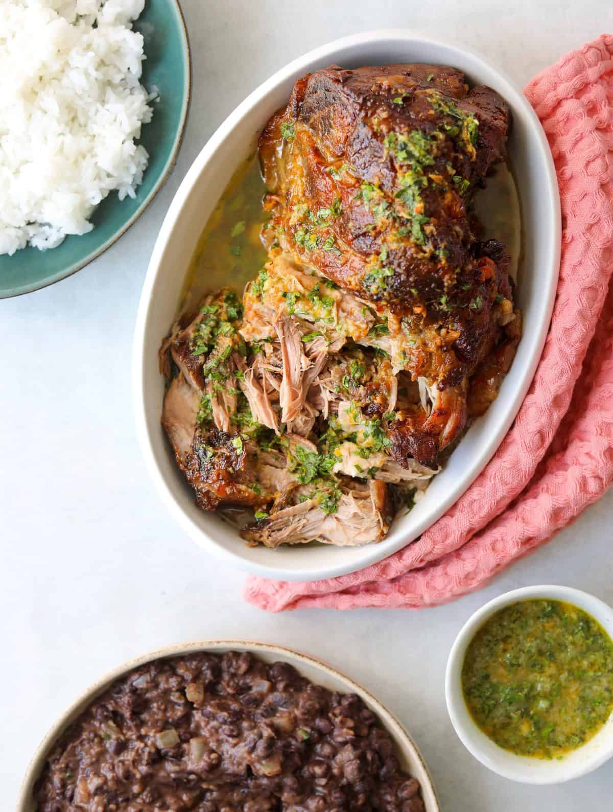 tender roasted pork in an oval baking dish with side dishes of rice, beans and mojo sauce