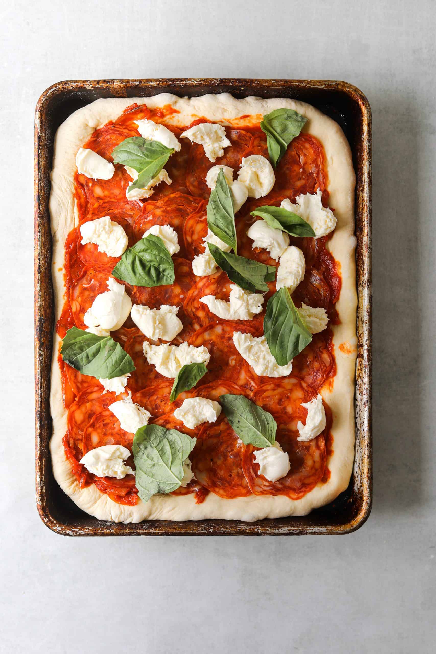 a rectangle pizza on a baking tray topped with soppressata, fresh mozzarella and basil 
