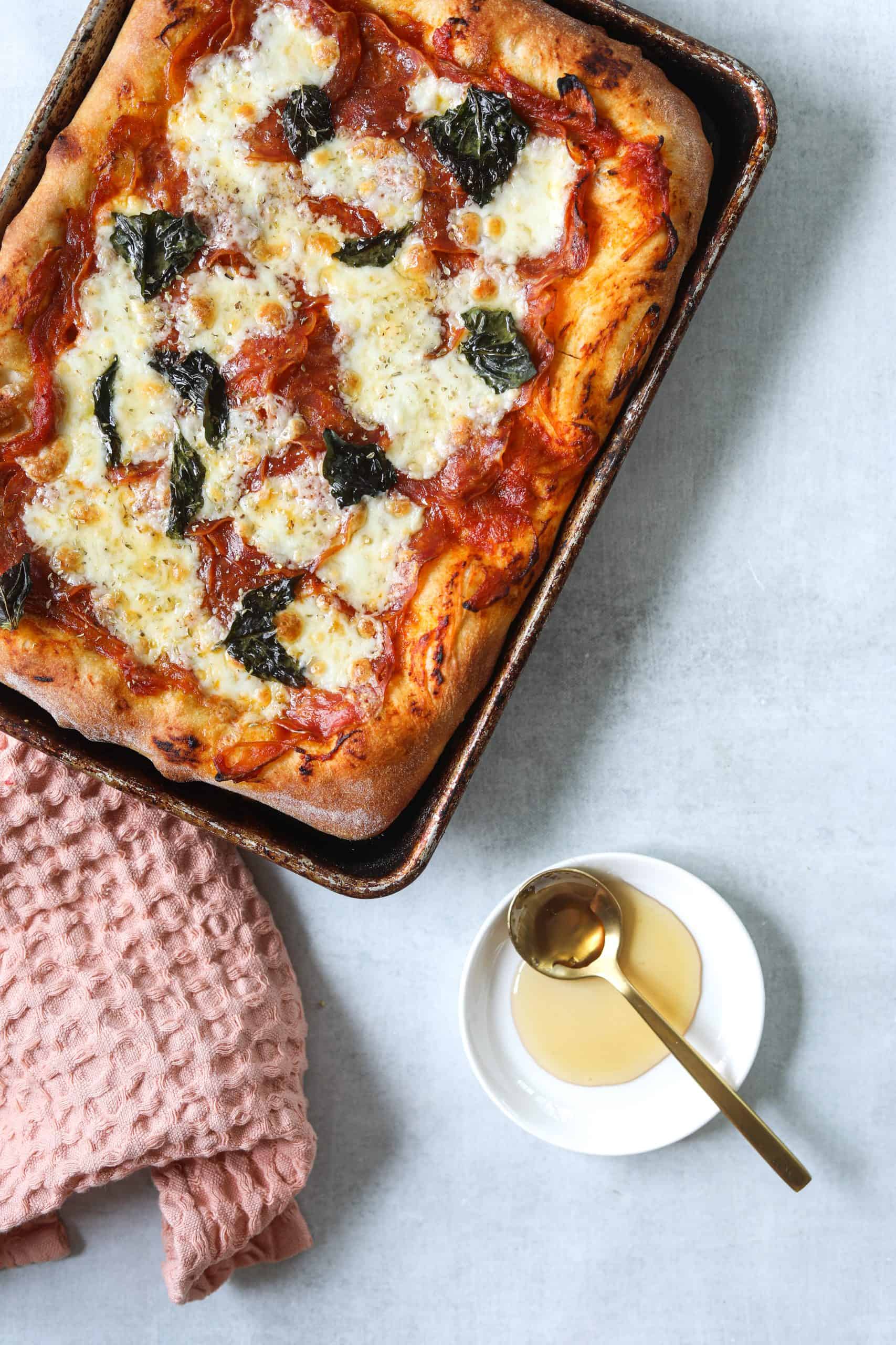 a rectangle pizza on a sheet pan with a side of honey and a pink tea towel