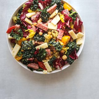 a white bowl of california chopped salad with provalone cheese, kale, tomatoes and salami on a blue background