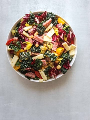 a white bowl of california chopped salad with provalone cheese, kale, tomatoes and salami on a blue background