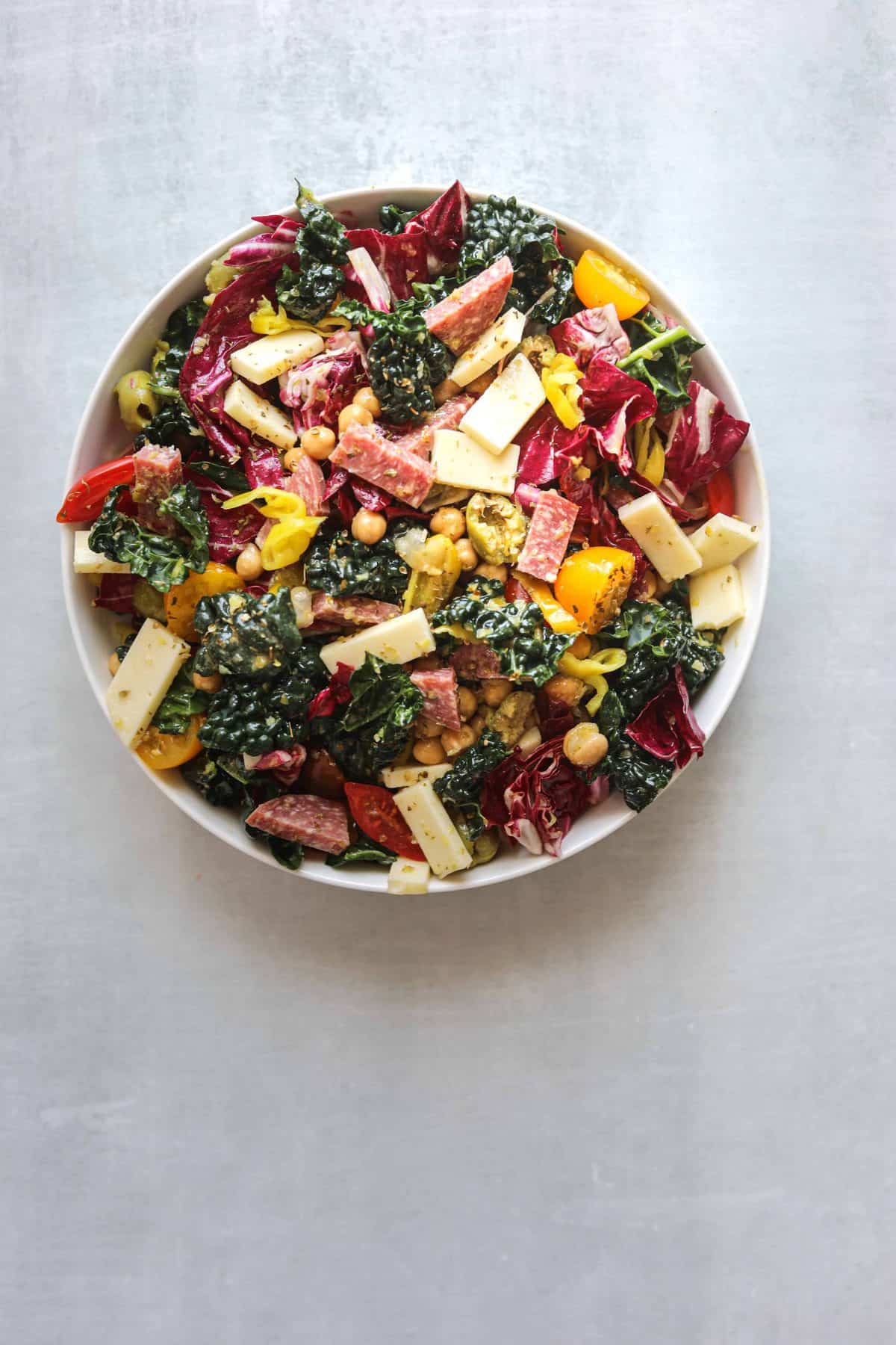 a white bowl of california chopped salad with provalone cheese, kale, tomatoes and salami on a blue background