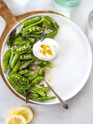 A white plate with snap pea and burrata salad, a silver spoon and two lemons