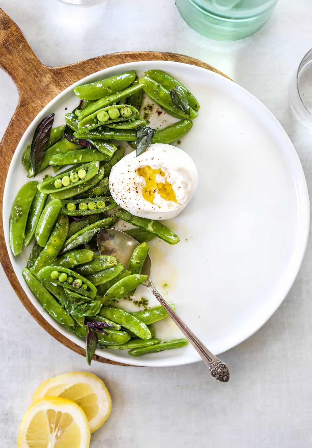 A white plate with snap pea and burrata salad, a silver spoon and two lemons