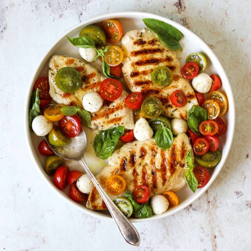 A white platter with three grilled chicken breasts topped with marinated tomatoes, fresh mozzarella cheese and basil.