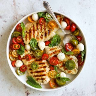 A white platter with three grilled chicken breasts topped with marinated tomatoes, fresh mozzarella cheese and basil.