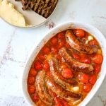 an oval baking dish filled with roasted sausages and tomatoes with grilled bread on the side
