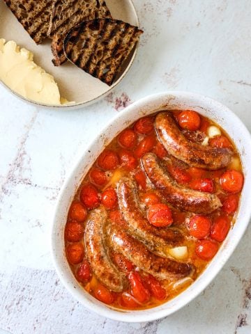 an oval baking dish filled with roasted sausages and tomatoes with grilled bread on the side