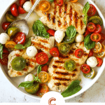 A white platter with three grilled chicken breasts topped with marinated tomatoes, fresh mozzarella cheese and basil.