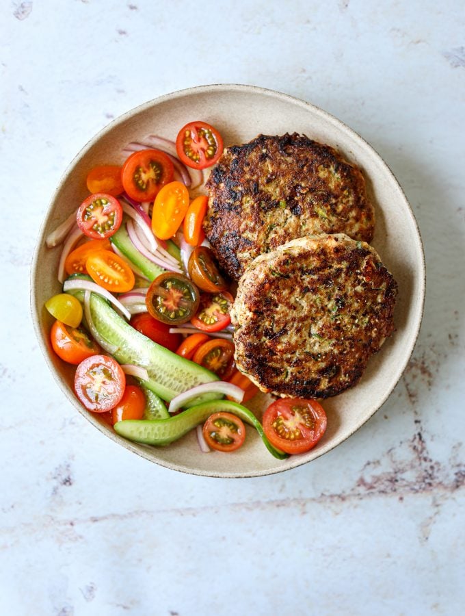 Two turkey and zucchini burgers on a plate with a side of tomato and cucumber salad