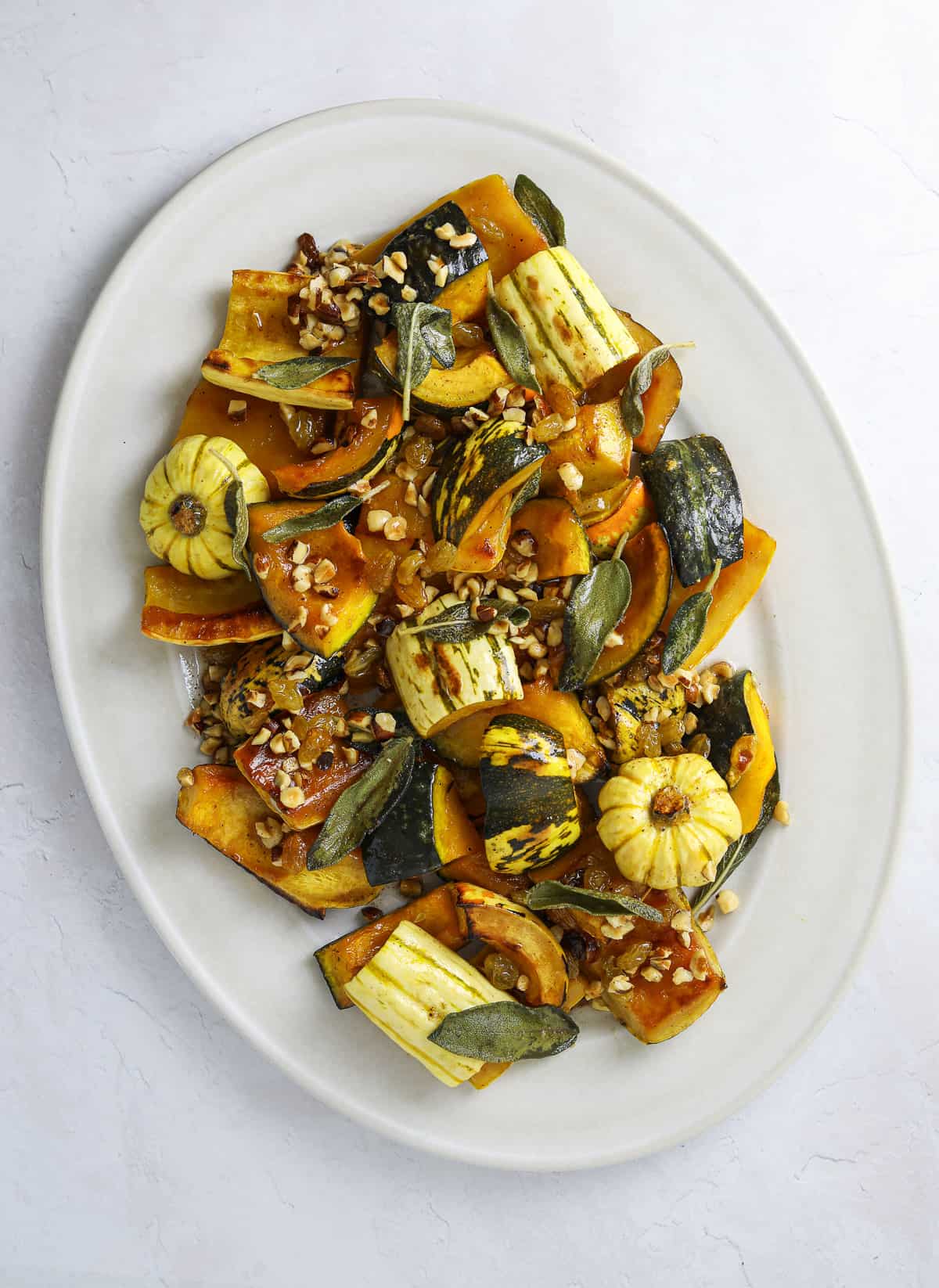 a white oval platter filled with roasted squash, sage leaves, hazelnuts and, raisins.