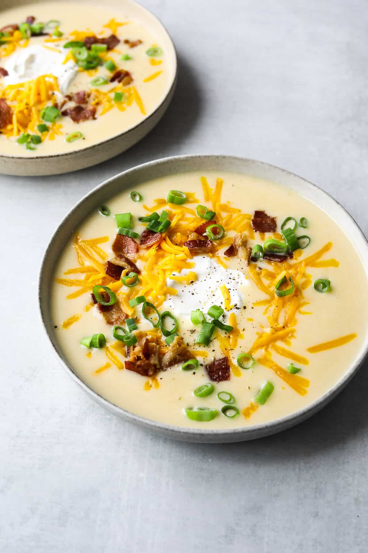 two bowls of cheesy potato soup topped with shredded cheddar cheese, crispy bacon bits and sliced green onion. 
