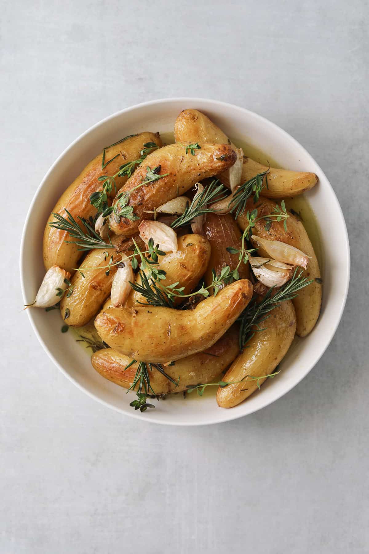 roasted fingerling potatoes, garlic, rosemary and thyme in a round white bowl on a blue background