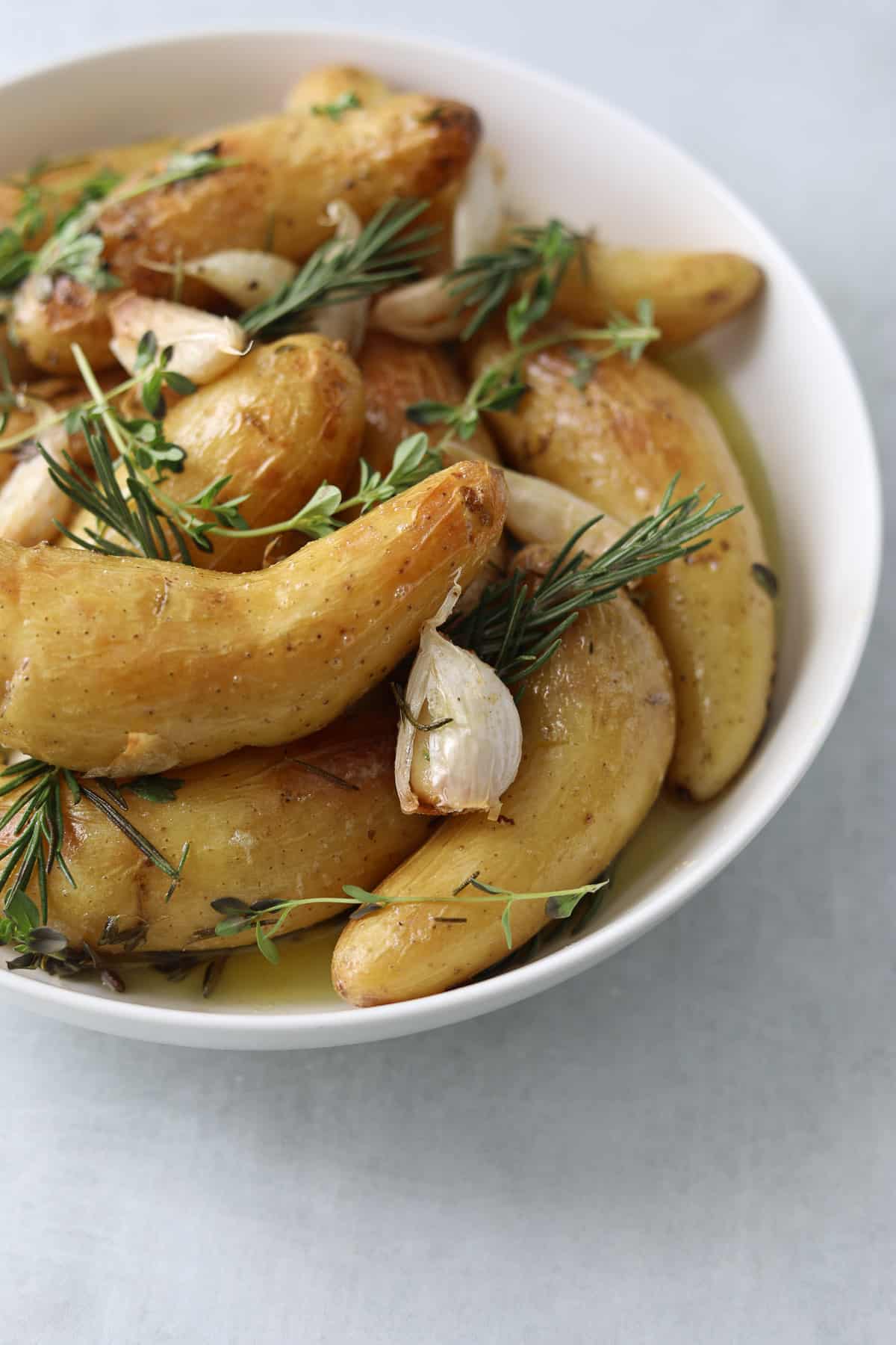 a white bowl filled with roasted  potatoes, herbs, garlic and oil on a blue background