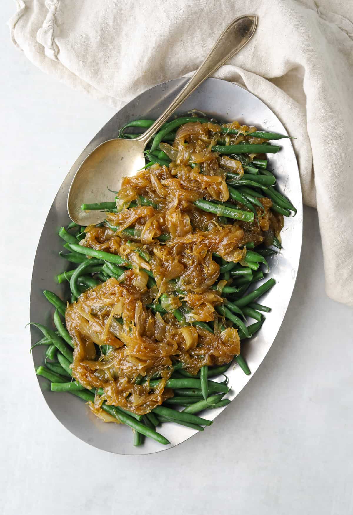 a silver platter filled with french green beans topped with golden brown caramelized onion next to a beige cloth napkin