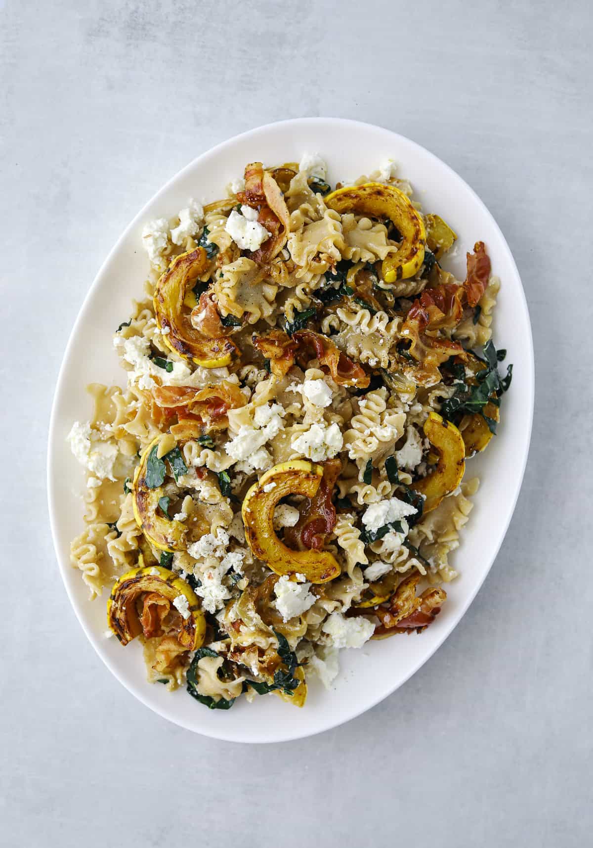 a white oval platter filled with pasta, goat cheese, roasted squash, and crispy pancetta