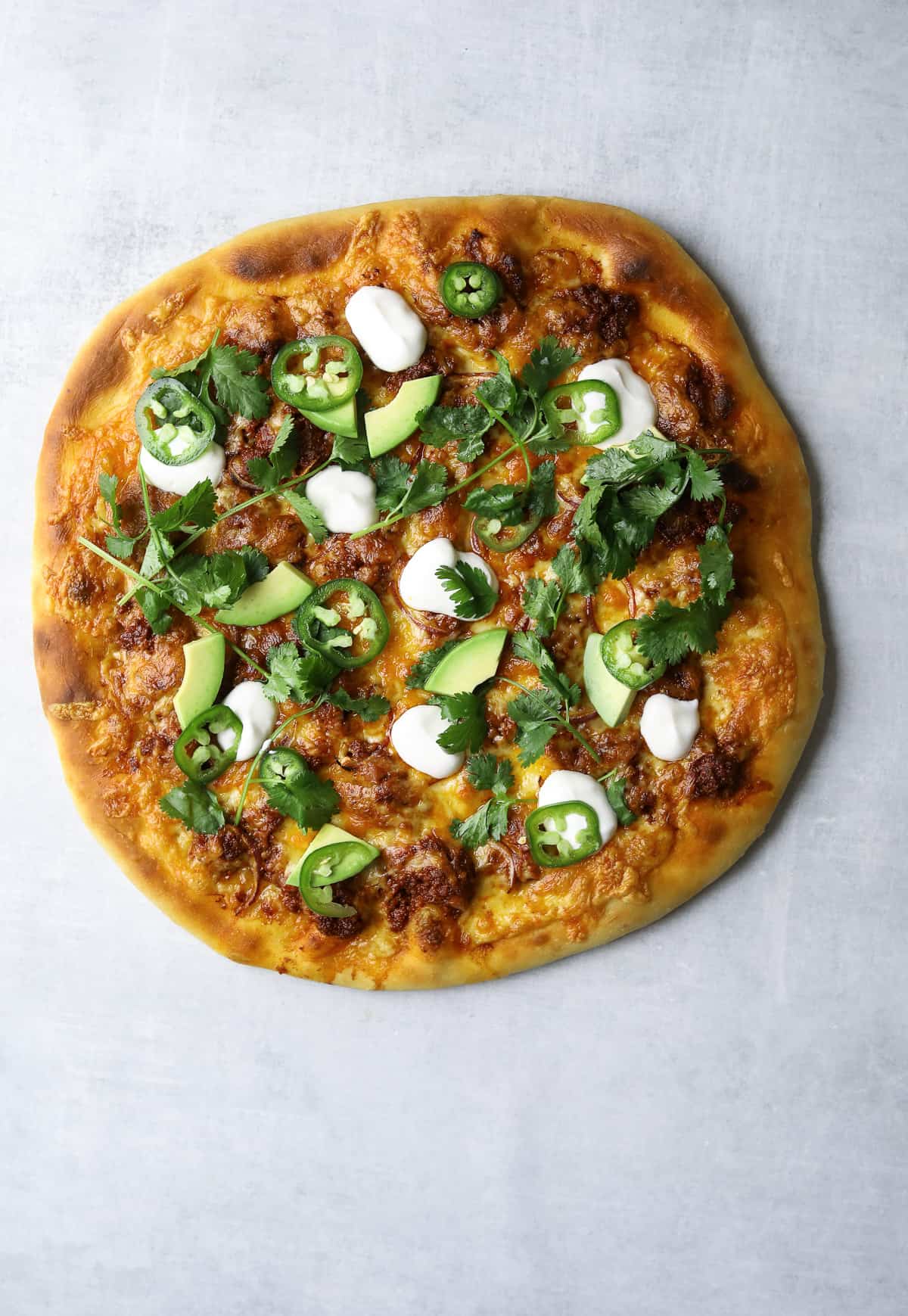 a pizza topped with chorizo, avocado, jalapeño slices, cilantro and sour cream on a blue background  
