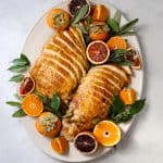 two roasted turkey breasts thinly sliced on an oval serving platter surrounded but citrus, herbs and persimmons