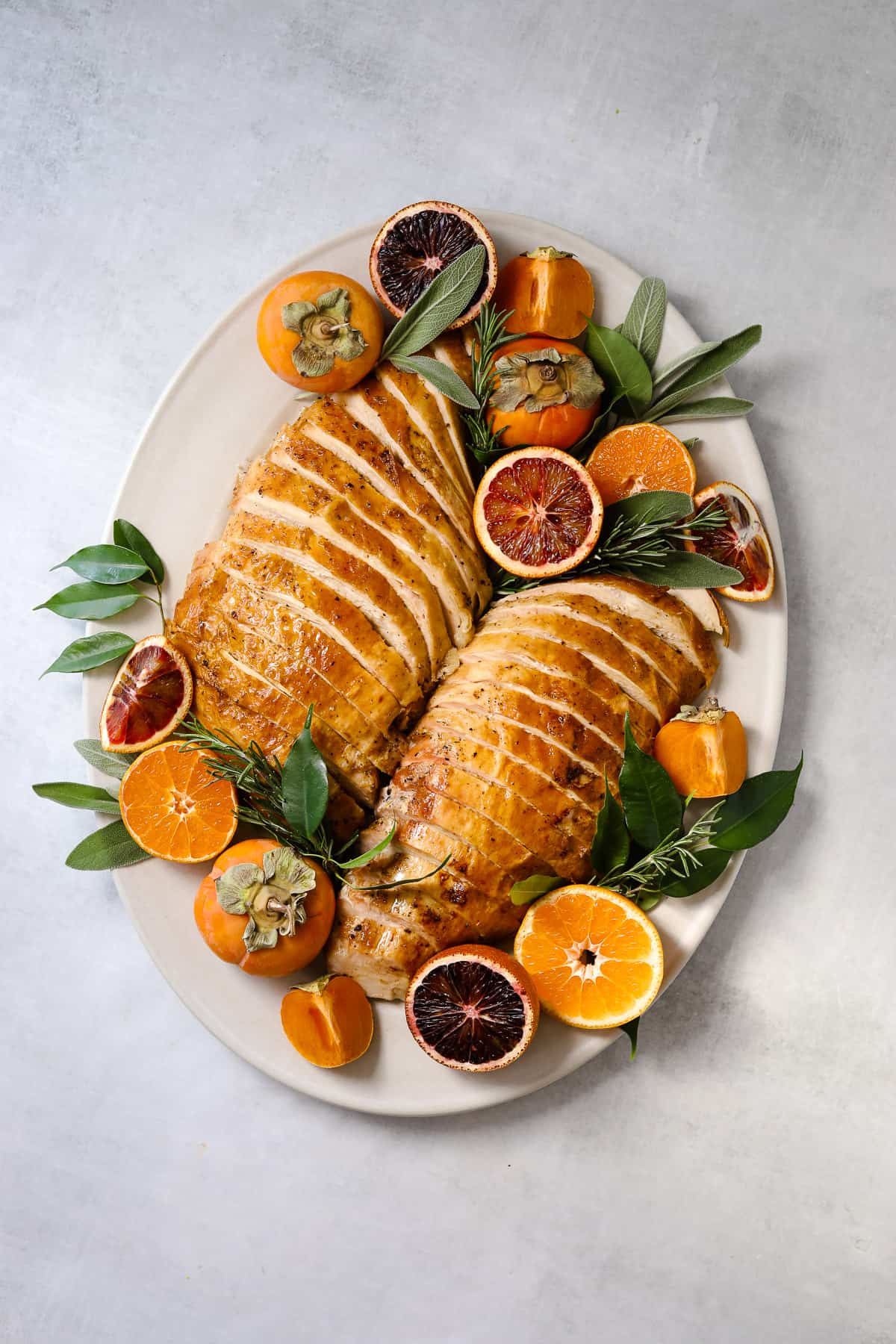 two roasted turkey breasts thinly sliced on an oval serving platter surrounded but citrus, herbs and persimmons 