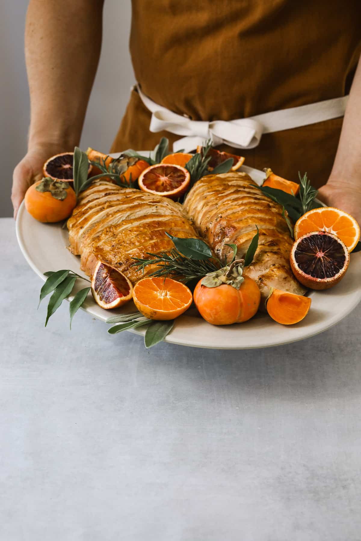 a person in a brown chefs apron presenting a platter of Roasted Turkey Breast garnished with fresh herbs, citrus and persimmons