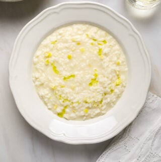 A white bowl filled with creamy risotto on a marble tabletop with white lace napkins and a glass of white wine.