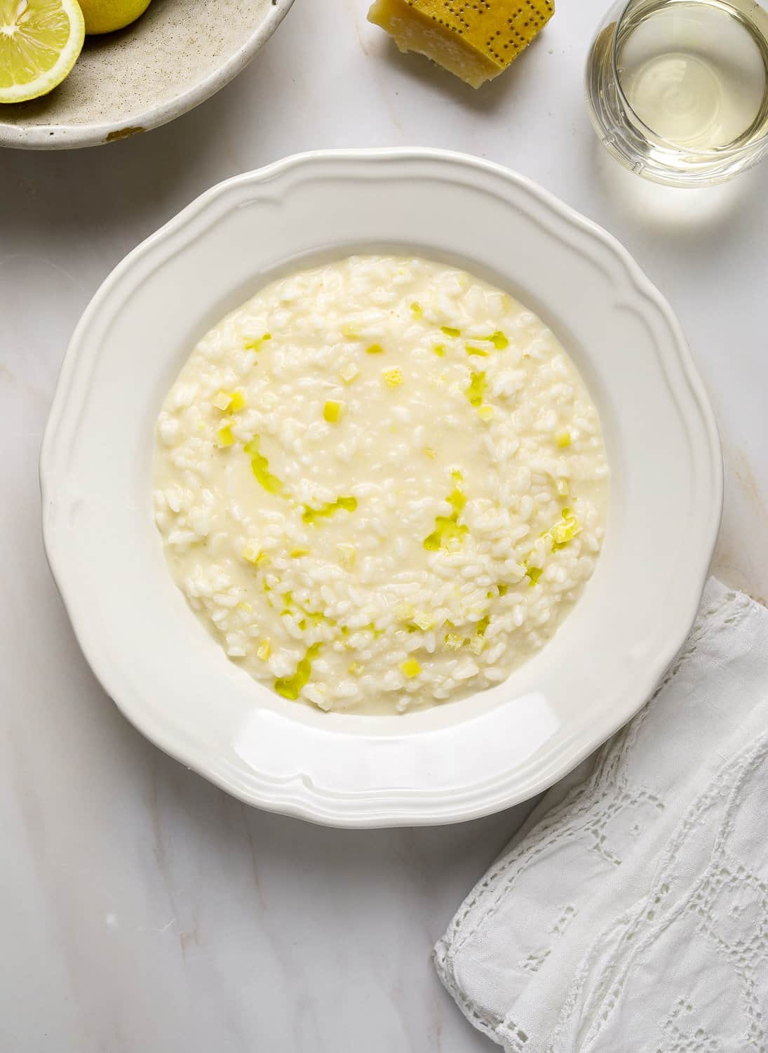 A white bowl filled with creamy risotto on a marble tabletop with white lace napkins and a glass of white wine.