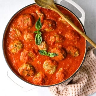 a white dutch oven skillet filled with baked meatballs, marinara sauce, fresh basil with a wooden spoon and pink towel