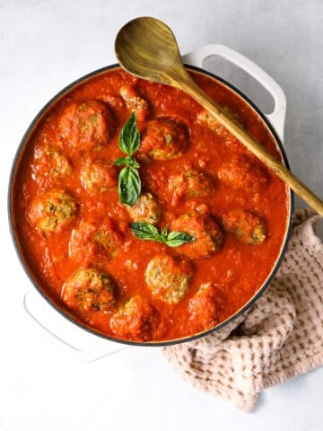 a white dutch oven skillet filled with baked meatballs, marinara sauce, fresh basil with a wooden spoon and pink towel