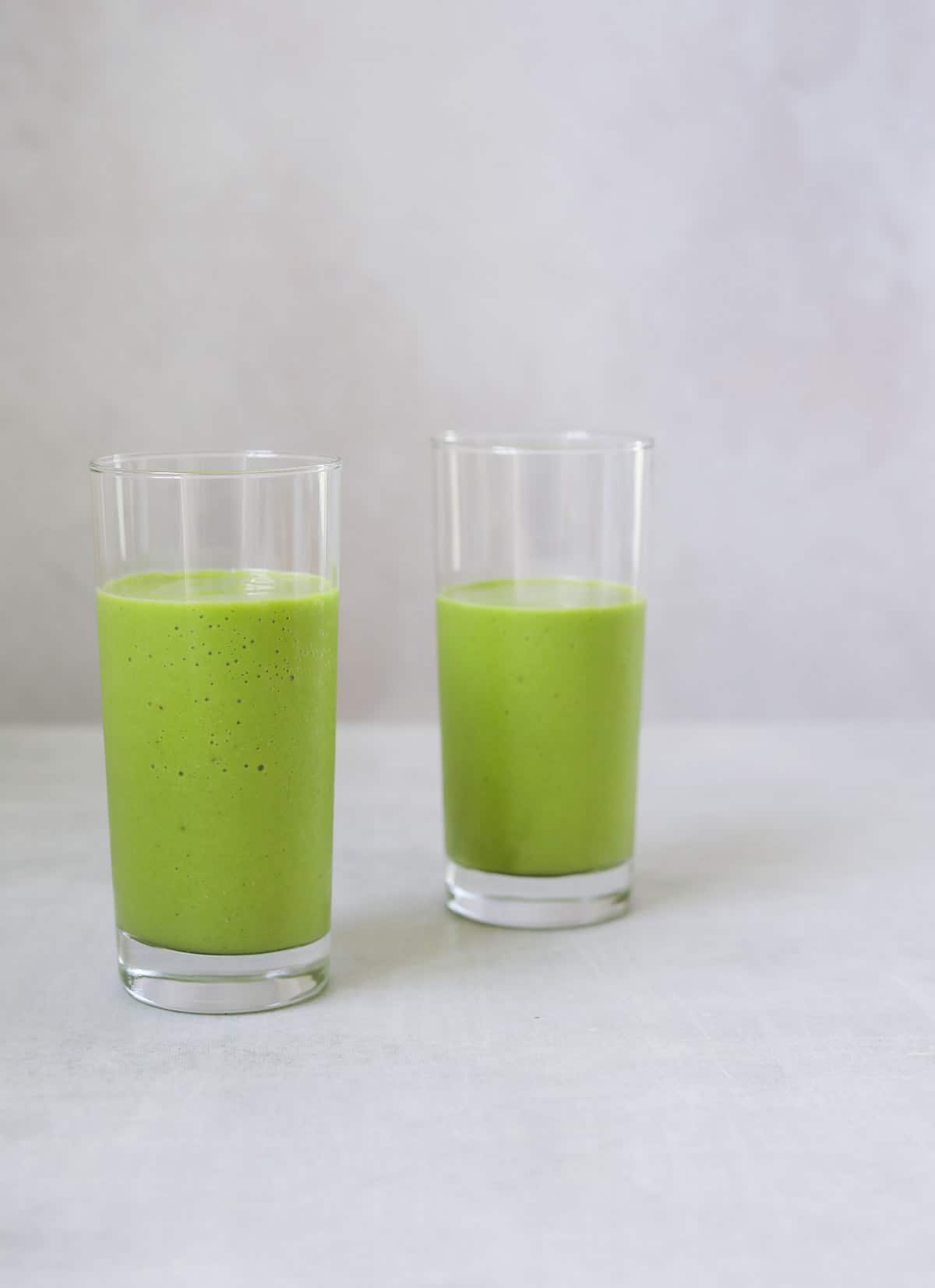 two glasses of bright green mango and pineapple smoothie on a blue background