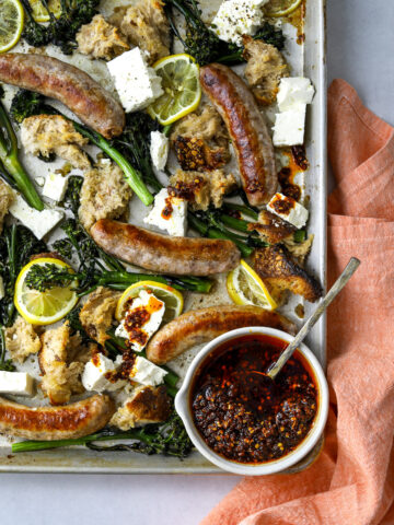 a sheet pan with roasted sausages, broccolini, lemon slices and bowl of chili oil