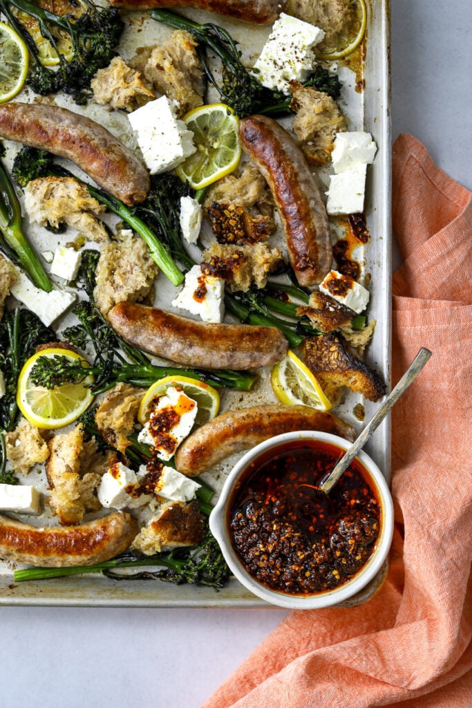 a sheet pan with roasted sausages, broccolini, lemon slices and bowl of chili oil