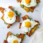 four open-faced breakfast sandwiches topped with arugula, bacon and sunny side up eggs.