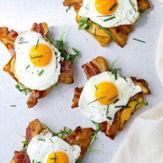 four open-faced breakfast sandwiches topped with arugula, bacon and sunny side up eggs.
