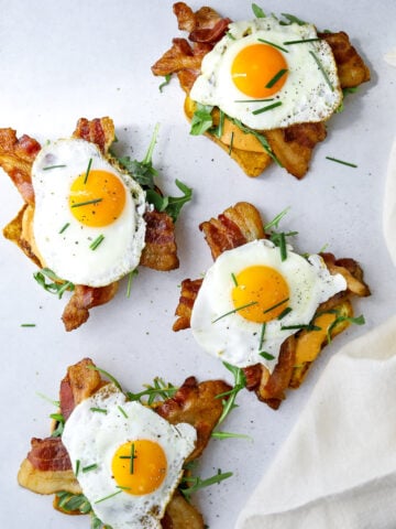 four open-faced breakfast sandwiches topped with arugula, bacon and sunny side up eggs.