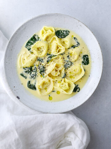 a ceramic bowl filled with a creamy soup of tortellini, spinach and topped with freshly cracked pepper and parmesan cheese