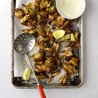 a paper towel lined baking tray with fried artichokes, lemon wedges and a bowl of lemon aioli