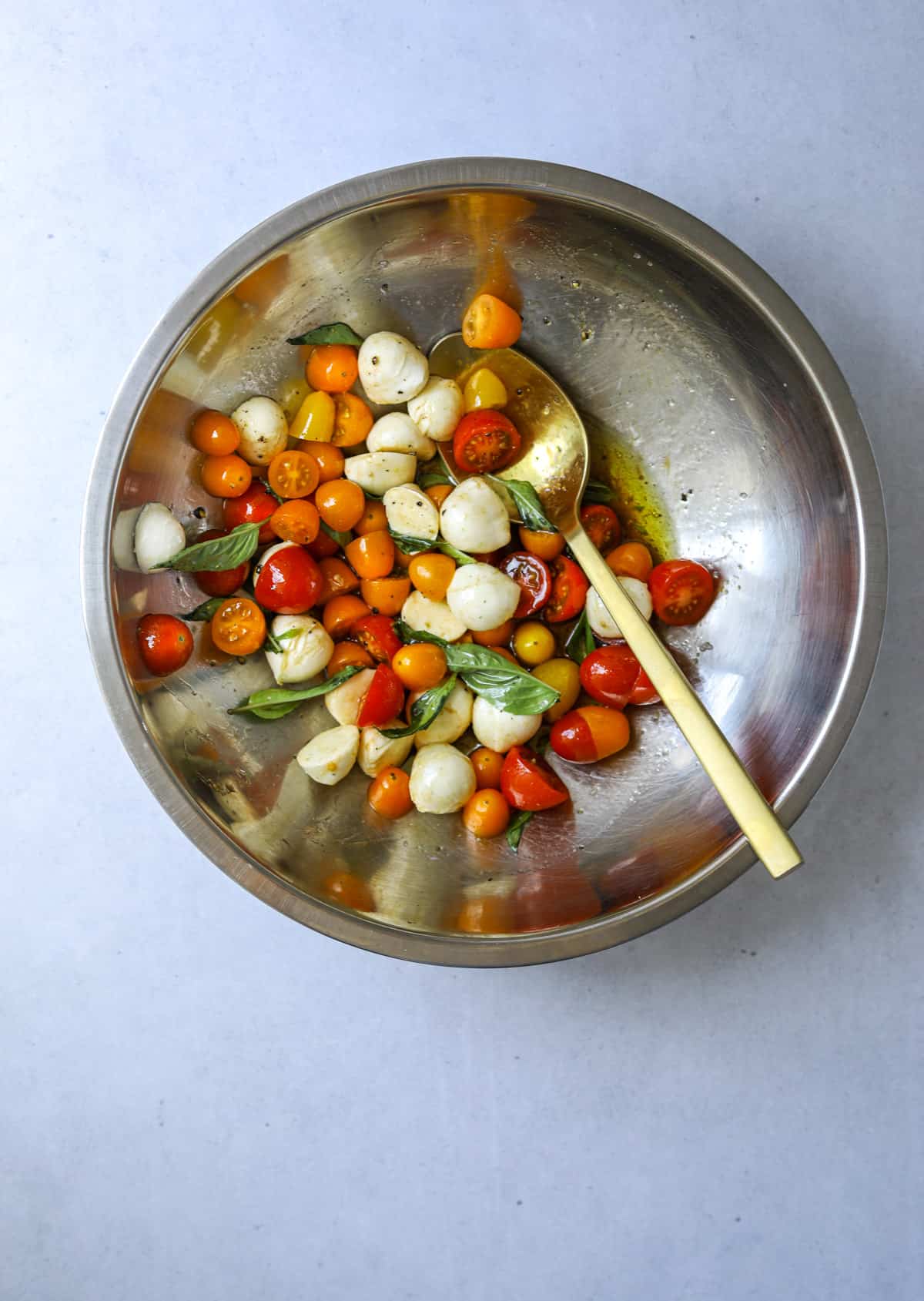 a stainless steel mixing bowl with cherry tomatoes, fresh mozzarella balls, fresh basil, olive oil and vinegar with a large gold mixing spoon