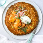 a white plate filled with a crispy, golden brown hash brown frittata topped with smoked salmon, a dollop of sour cream and fresh herbs. blue knife on the side, white background.