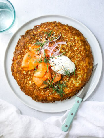 a white plate filled with a crispy, golden brown hash brown frittata topped with smoked salmon, a dollop of sour cream and fresh herbs. blue knife on the side, white background.