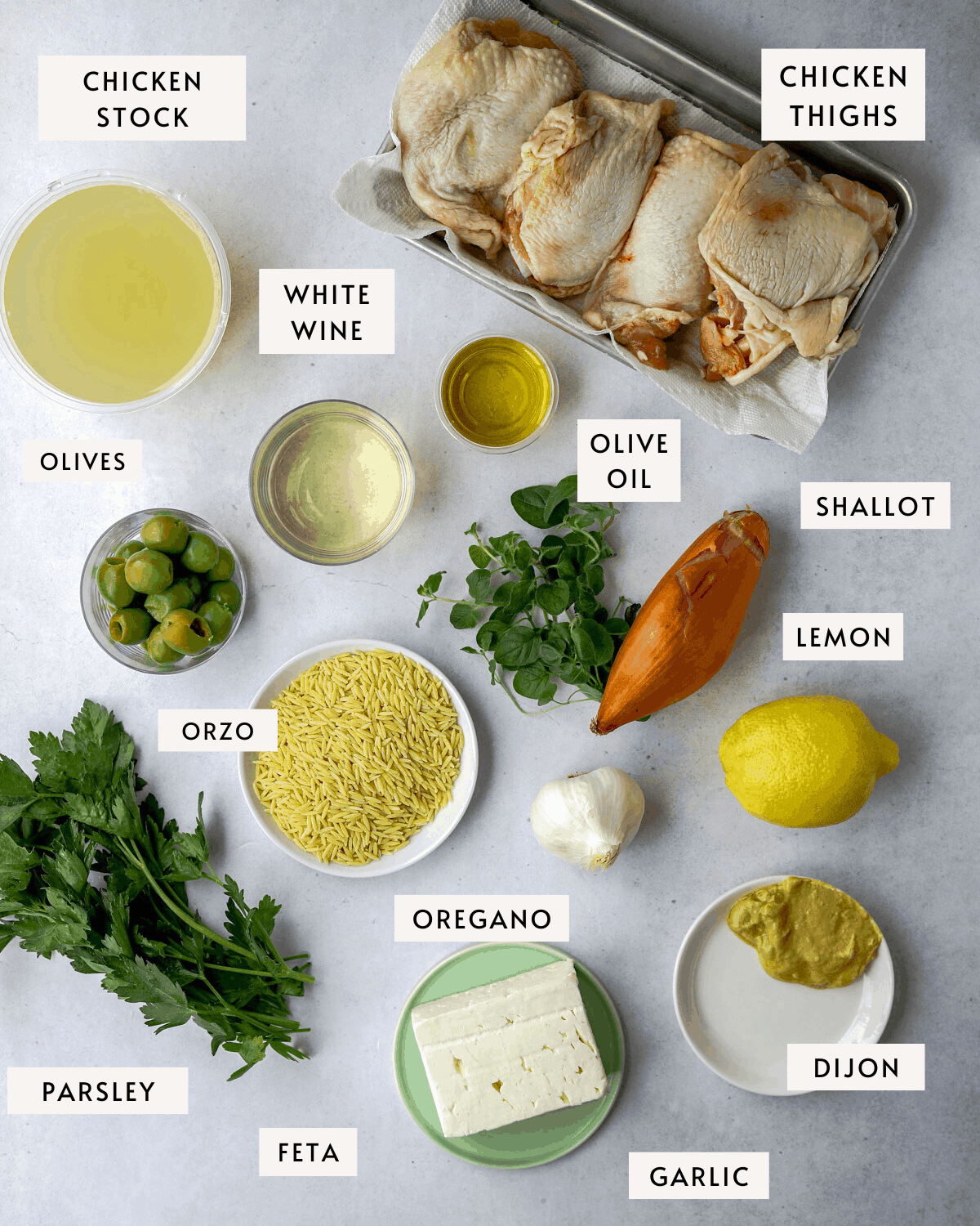 recipe ingredients individually portioned on a blue background; orzo, chicken thighs, feta cheese, lemons, garlic, olive oil, white wine, olives, and herbs