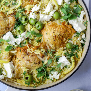 a pot of orzo and chicken thighs topped with feta, olives, lemons and fresh herbs on a blue background