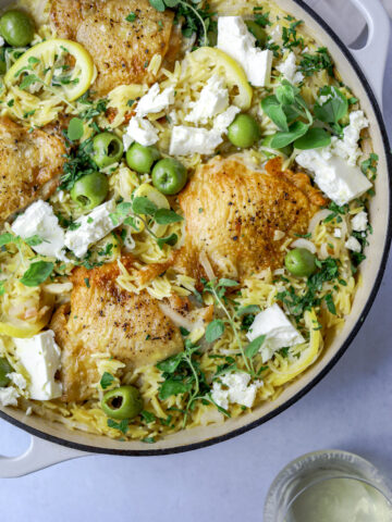 a pot of orzo and chicken thighs topped with feta, olives, lemons and fresh herbs on a blue background