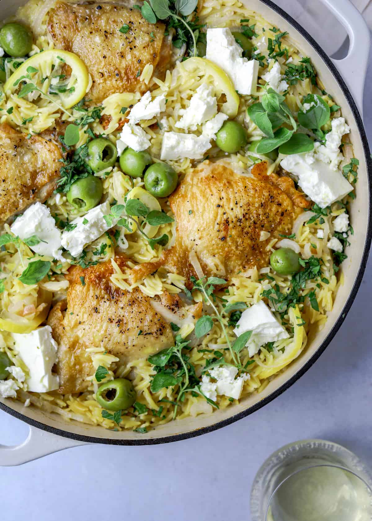 a pot of orzo and chicken thighs topped with feta, olives, lemons and fresh herbs on a blue background