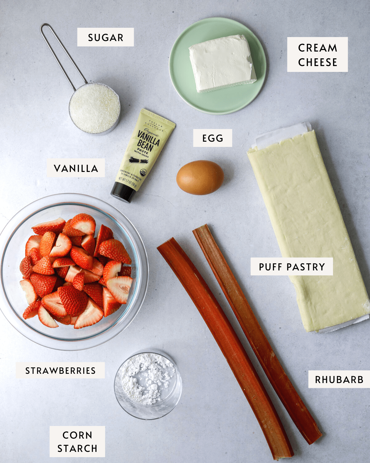 recipe ingredients: frozen puff pastry, cream cheese, vanilla, a bowl of fresh strawberries, two stalks of rhubarb and a measuring cup of sugar