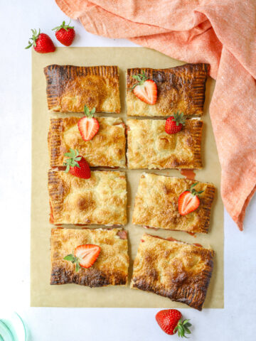 a strawberry rhubarb puff pastry tart cut into 8 rectangles on a blue background with fresh strawberries on top and a pink cloth napkin